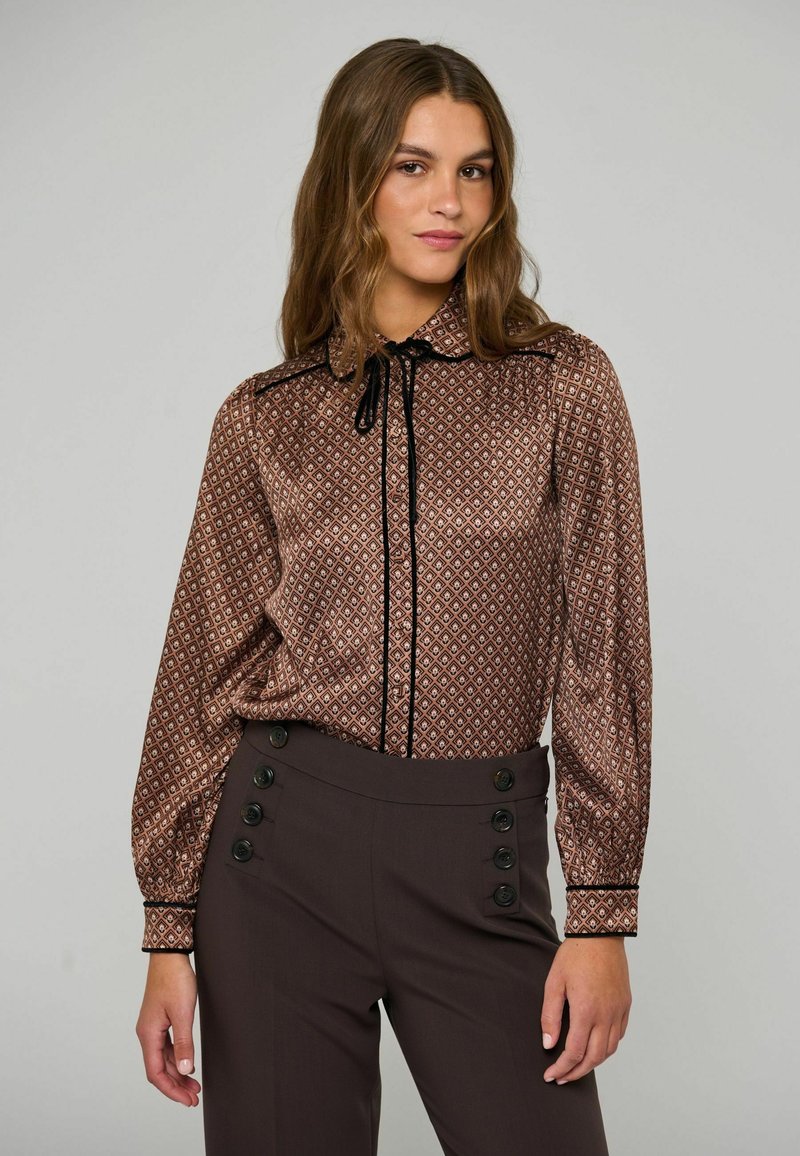 Jeune femme aux longs cheveux porte un chemisier marron à motifs avec bordure noire et un pantalon sombre taille haute avec des boutons décoratifs.