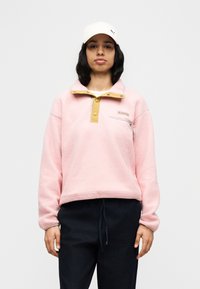 Ροζ πουλόβερ από φλίς με σχεδιασμό με φερμουάρ στο ένα τέταρτο, κίτρινο γιακά και τσέπη στο στήθος. Φορεμένο με σκούρο παντελόνι με κορδόνι. Μαλακή υφή συνολικά.