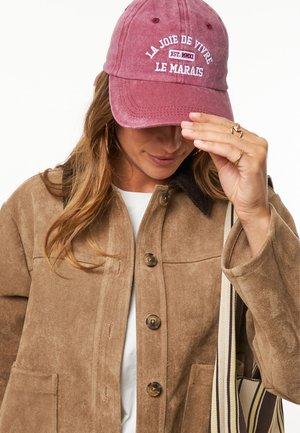 Bruine suède jas met een corduroy kraag en knoopsluiting, gecombineerd met een bordeauxkleurige pet met geborduurde tekst. Een wit shirt is zichtbaar eronder.