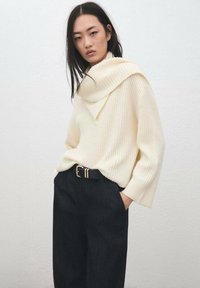 Pull en maille côtelée crème avec un large col rabattu, associé à un pantalon en denim foncé taille haute et une ceinture noire ornée d'accents dorés.