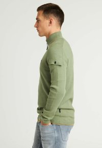 Grüner Strickpullover mit hohem Kragen, ausgestattet mit einem Reißverschluss und schwarzen Akzenten, ergänzt durch hochgerollte Ärmel und eine enganliegende Passform.