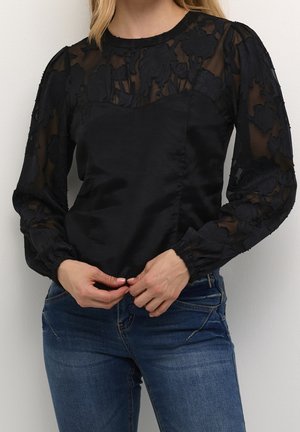 Frau trägt eine schwarze, durchsichtige, langärmlige Bluse mit floraler Stickerei, kombiniert mit blauen Jeans, die Hände vor der Taille verschränkt.