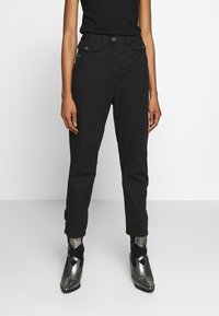 Pantalones cargo negros de corte ajustado, con bolsillos frontales y cierre de botón. Combinados con botas de tobillo metálicas, sobre un fondo blanco liso.