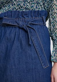 Jupe en denim avec une taille froncée, ceinture nouée, couleur bleu foncé, et détails de couture visibles. Portée avec un haut à motif floral.