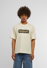 Lys beige bomulls t-skjorte med løs passform, som har et grønt brodert "MARKET?" design omkranset av fargerike blomsterdetaljer.