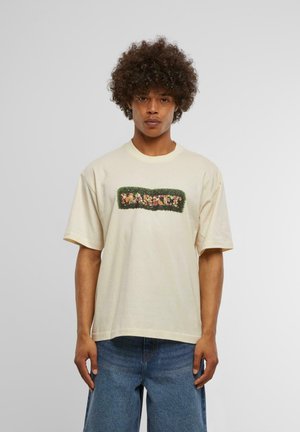 Lyse beige bomulds t-shirt i løs pasform med et grønt broderet "MARKET?" design omgivet af farverige blomsterdetaljer.