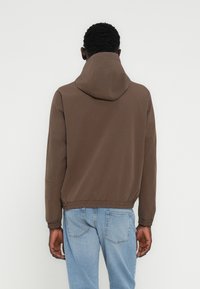 Καφέ μπουφάν με κουκούλα και σφιχτή μέση, μινιμαλιστικός σχεδιασμός, ομαλή υφή, φορεμένο πάνω από ανοιχτόχρωμα μπλε τζιν, θέα από πίσω.