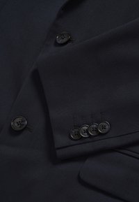 Sötétkék blézer sima anyagból, két nagy fekete gombbal és négy kisebb gombbal, kontrasztos varrással a mandzsettán.