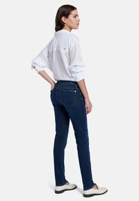 Chemise blanche à boutons avec des poignets retroussés, associée à un jean skinny bleu foncé. Détail remarquable dans le dos avec une étoile jaune. Chaussures décontractées.
