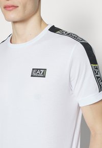 T-shirt en coton blanc avec une bande graphique noire et néon sur l'épaule et un petit logo EA7 sur la poitrine. Manches courtes.