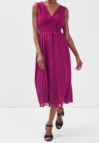 Robe plissée fuchsia avec un décolleté en V et de larges bretelles. Détail froncé à la taille, longueur mi-mollet, portée avec des talons noirs à bride cheville.