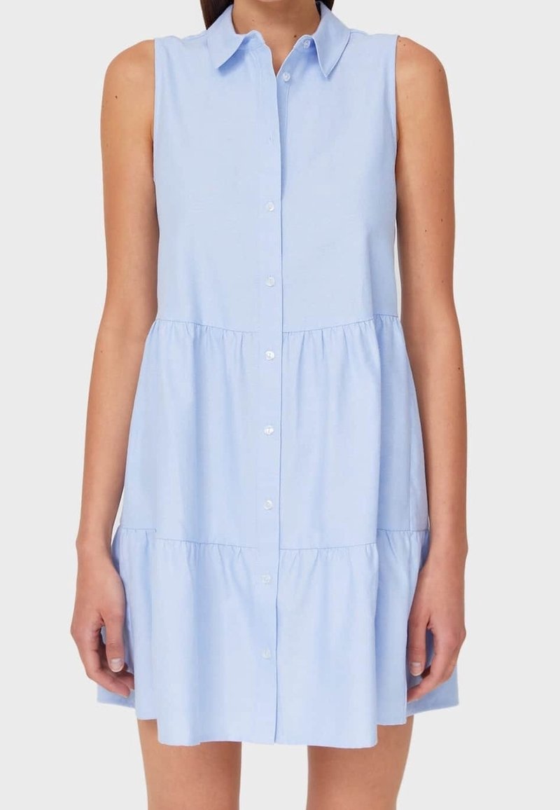 Robe sans manches bleu clair à boutons, avec col et jupe froncée à volants portée par une personne debout sur un fond uni.