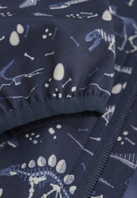 Marinblått tyg med vita dinosaurieskelett, ägg och ben, med en rynkad kant och dragkedjestängning. Mjuk textur, lekfull design.