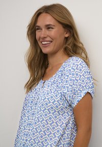 Blouse à motifs bleus et blancs avec des manches courtes, un col rond et un détail en trou de serrure à l'avant, fabriquée dans un tissu doux et texturé.