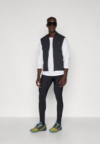 Schwarze ärmellose Weste über einem weißen Langarmshirt, kombiniert mit schwarzen Leggings und grünlich-gelben Laufschuhen mit blauen Akzenten.