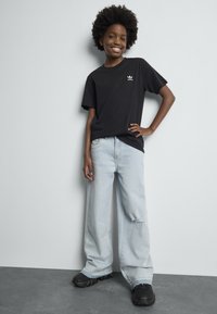 Smilende ung person med afrofrisyre som har på seg en svart Adidas t-skjorte, lyseblå vide jeans med hull, og svarte joggesko, som står mot en vegg.