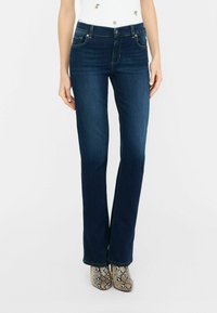Jeans acampanados de color azul oscuro, hechos de denim con una textura suave, que cuentan con bolsillos frontales y cierre de cremallera, combinados con zapatos de serpiente metálica.
