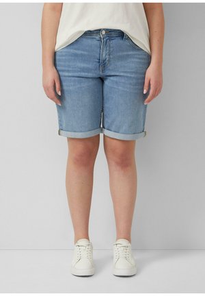 Person trägt hochgekrempelte blaue Jeansshorts, weiße Sneakers und ein weißes Oberteil und steht vor einem schlichten grauen Hintergrund.