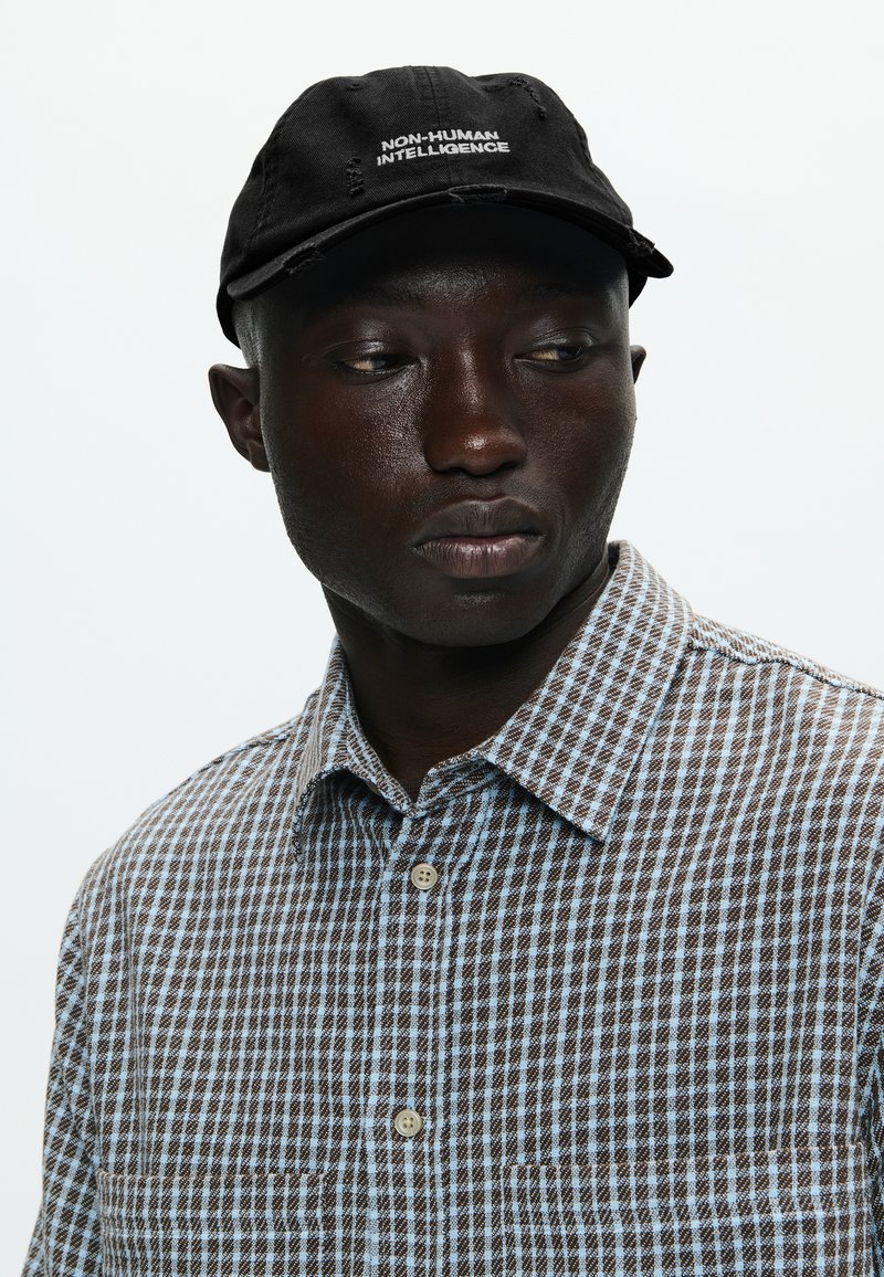 Zwarte pet met de tekst "NON-HUMAN INTELLIGENCE", gecombineerd met een bruine en lichtblauwe geblokte shirt met een knoopsluiting en een zak.
