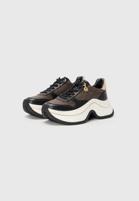 Bruine en zwarte chunky sneakers met dikke witte zolen, een gouden logo charm en glanzende beige hieldetails op een effen lichte achtergrond.