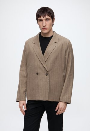 Homme portant un blazer beige à fines rayures et à double boutonnage sur une chemise noire, debout devant un fond blanc uni.
