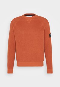 Stickad orange tröja med rund halsringning, ribbade ärmslut och nederkant, samt ett litet svart logotypmärke på vänster ärm.