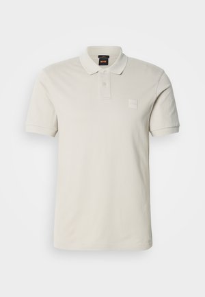 Polo de color beige claro hecho de algodón suave, con cuello clásico, mangas cortas, dos botones y un pequeño parche con logo en el pecho.