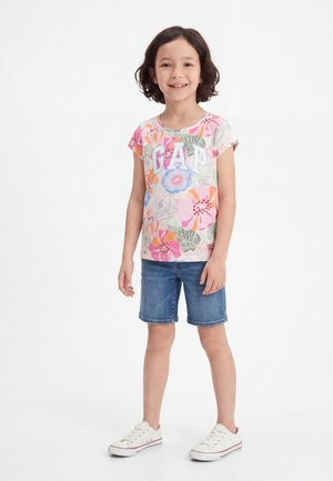 Χαμογελαστό κορίτσι φοράει πολύχρωμο φλοράλ μπλουζάκι GAP, τζιν σορτσάκι και άσπρα αθλητικά, στέκεται μπροστά σε λευκό φόντο.