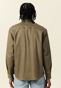 Camicia a maniche lunghe verde oliva con taglio dritto, polsini abbottonati e dettaglio della yoke posteriore. Si abbina a jeans azzurri chiari.