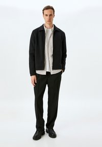 Chaqueta negra con cremallera y cuello, superpuesta a una camisa blanca con rayas rojas verticales. Combinada con pantalones y zapatos negros.