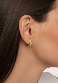 Pendiente de aro geométrico dorado con forma hexagonal y acabado pulido, llevado en la oreja. La vista en primer plano destaca su diseño elegante y color.