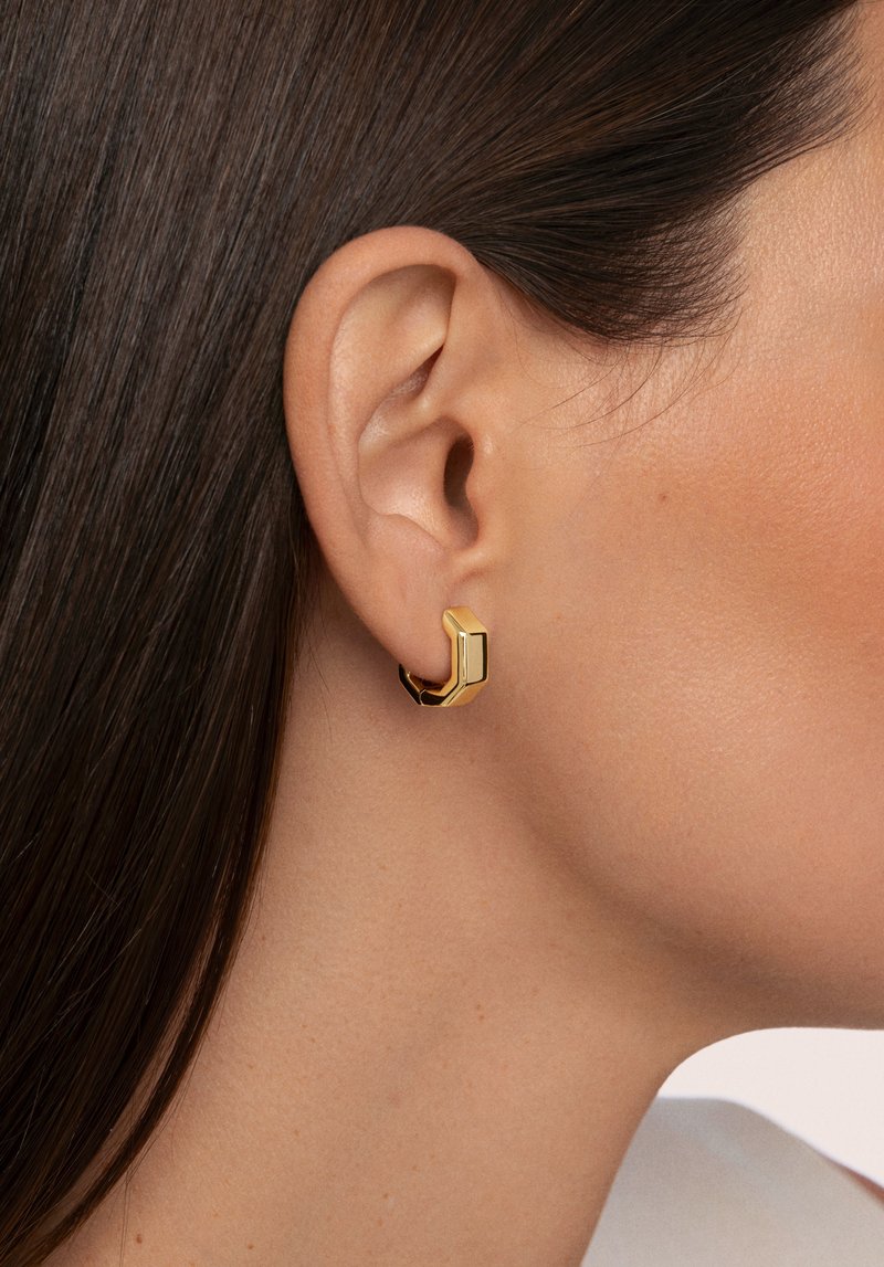 Pendiente de aro geométrico dorado con forma hexagonal y acabado pulido, llevado en la oreja. La vista en primer plano destaca su diseño elegante y color.