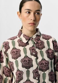 Hellbeige Hemd mit Kragen, verziert mit einem floralen Muster in Dunkelpink und grünen Blättern. Es hat Knöpfe und einen strukturierten Stoff.