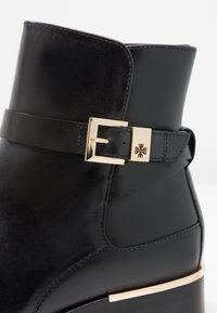 Bottine en cuir noir avec une boucle rectangulaire dorée et un accent doré assorti sur le bord du talon.