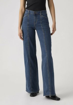 Mujer con vaqueros de pierna ancha azul de talle alto con costuras verticales, camiseta negra y zapatos negros de punta afilada sobre un fondo liso.