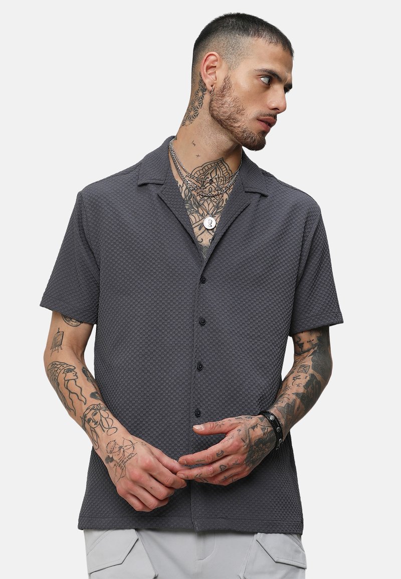 Camisa de manga corta, color gris oscuro, con un patrón texturizado, con botones en la parte delantera, cuello y detalles de costura visibles; llevada con collares en capas.