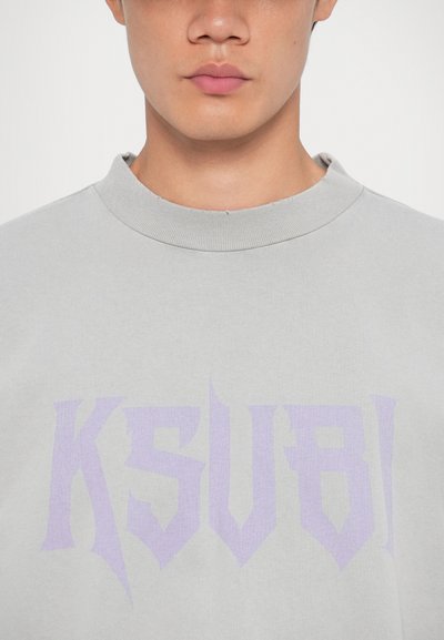 Lys grå hettegenser med høy krage, med et stort, stilisert lilla logo "KSUBI" på forsiden. Mykt, teksturert stoff.