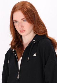 Zwarte zip-up hoodie met grijze koordjes, voorzien van een klein wit schiplogo op de borst en een zachte, gestructureerde stof.