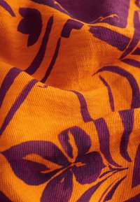Gros plan d’un tissu orange texturé avec un motif floral et abstrait violet, montrant les plis et les détails du tissage textile.