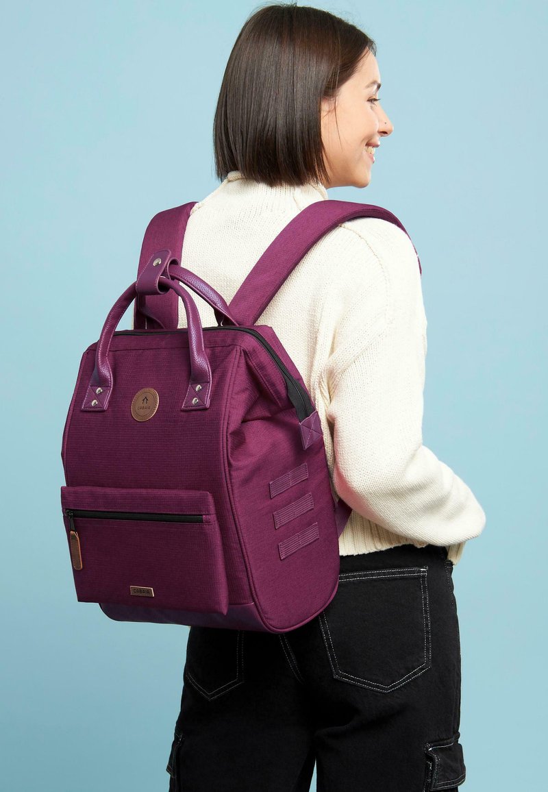 Zaino in tessuto viola con doppie maniglie superiori, spallacci regolabili, tasca frontale con zip e dettagli testurizzati. Design pulito senza loghi visibili.