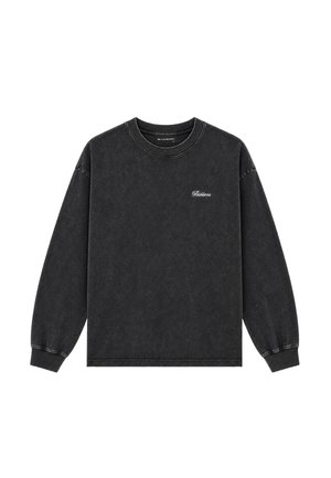 Mann steht und trägt ein schwarzes Langarmshirt, ausgebleichte gelb-grüne Jeans und schwarz-weiße Turnschuhe auf einem schlichten hellgrauen Hintergrund.