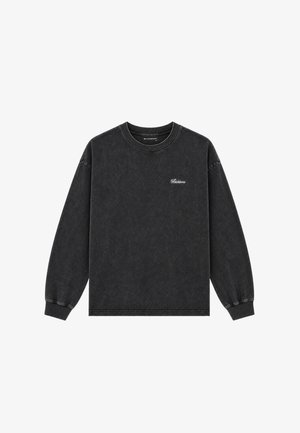 Mann steht und trägt ein schwarzes Langarmshirt, ausgebleichte gelb-grüne Jeans und schwarz-weiße Turnschuhe auf einem schlichten hellgrauen Hintergrund.