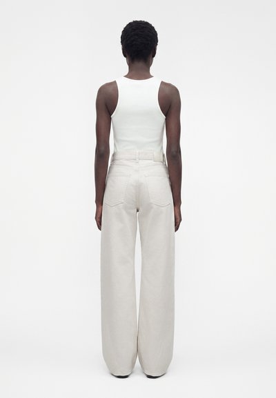 Femme debout de dos, vêtue d'un haut blanc sans manches et d'un pantalon beige taille haute à jambes larges, sur un fond uni.
