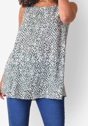 Femme portant un haut tunique sans manches noir et blanc à pois, associé à un jean bleu, tenant un bras près de sa tête.