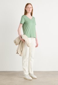Grønn v-hals t-skjorte laget av mykt stoff, kombinert med kremfargede bukser og hvite sneakers. En beige jakke er lagt over armen.