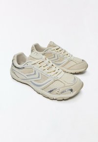 Paire de chaussures de course légères en mesh beige avec lacets blancs et motifs chevrons gris sur les côtés sur fond blanc.