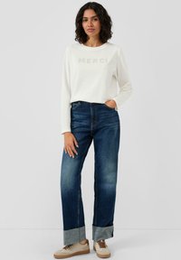 Biała koszula z długim rękawem z napisem "MERCI", zestawiona z niebieskimi dżinsami z podwiniętymi mankietami i jasnymi akcentami. Beżowe trampki.