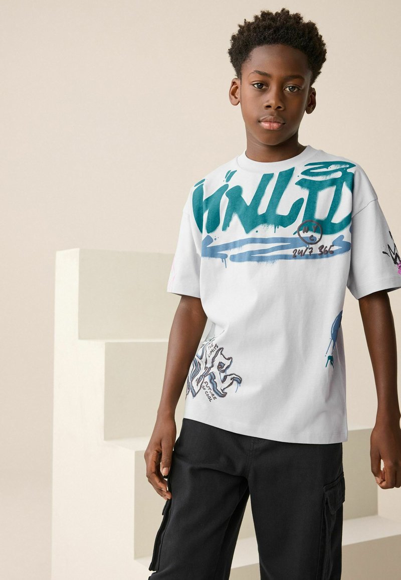 T-shirt bianco in cotone con grafiche in stile graffiti blu e verde, maniche corte e vestibilità casual. Abbinato a pantaloni cargo neri.