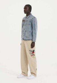 Denim-paita, jossa on kaksi etutaskua ja brodeerattu kuvio, yhdistettynä beigeihin leveisiin housuihin, joissa on värikkäitä tattoo-tyylisiä taideteoksia. Valkoiset tennarit.