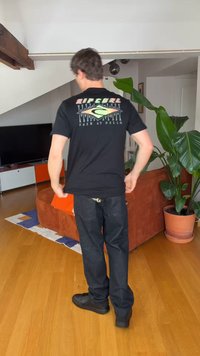 Férfi fekete Rip Curl pólóban és sötét farmernadrágban áll egy nappaliban, ahol fa padló, narancssárga kanapé, nagy zöld növény és TV szekrény látható.